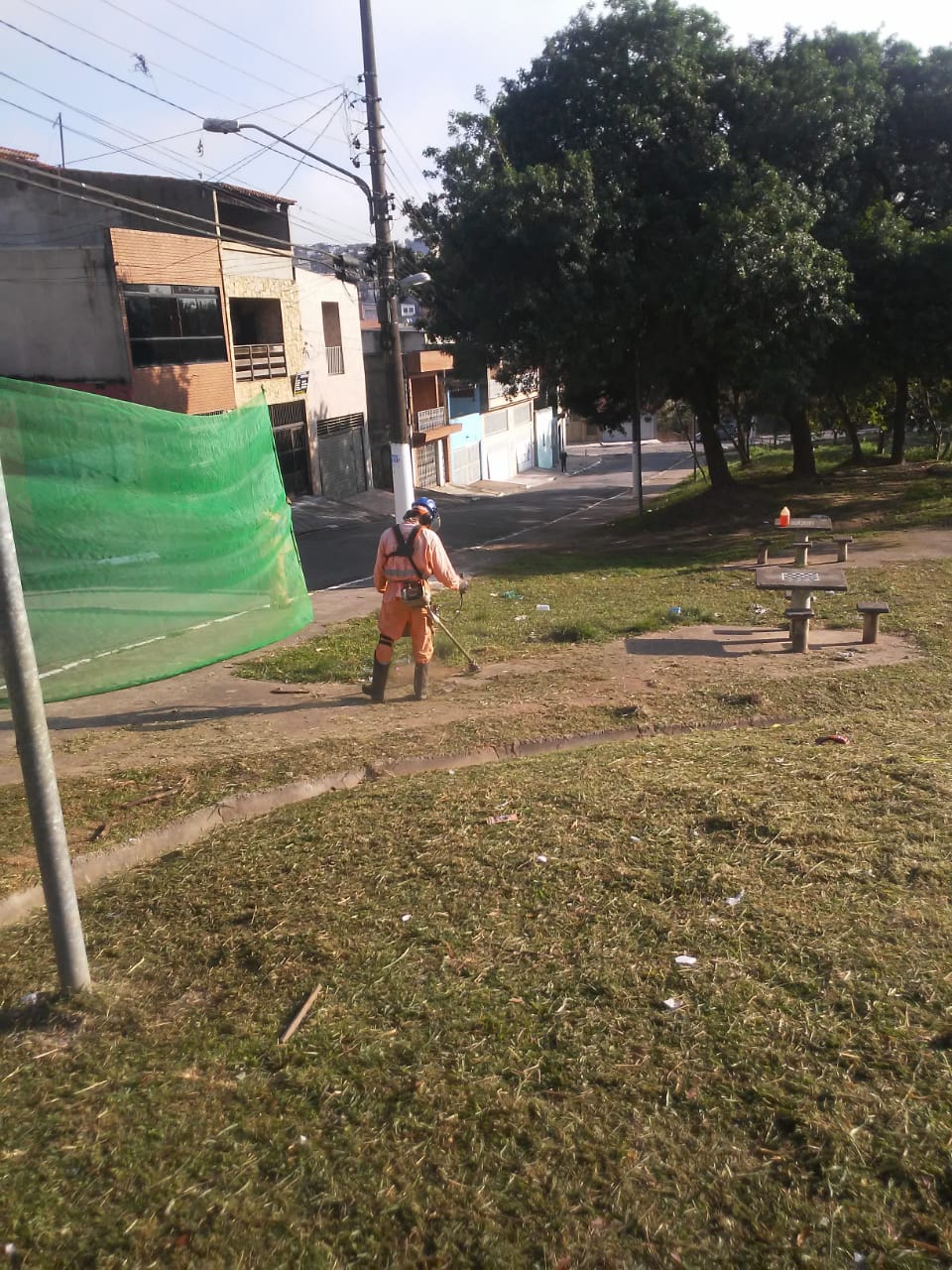 Funcionário com uniforme da Prefeitura podando grama com roçadeira; uma tela de proteção é vista à esquerda.